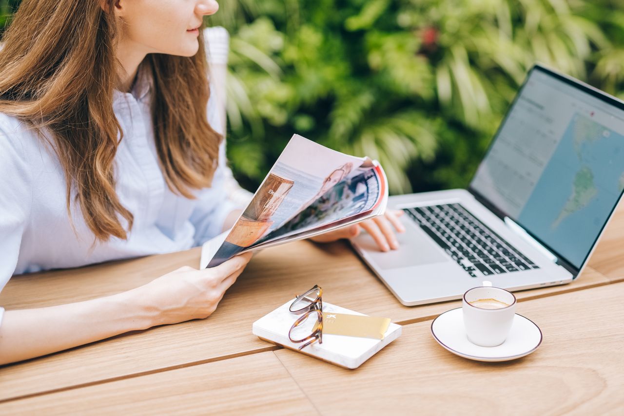 A woman sits outdoors, enjoying a magazine with coffee and laptop nearby.
